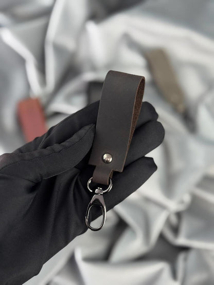 A dark brown leather keychain loop with a silver metal snap and key ring, held by a hand wearing a black glove against a draped silver fabric background.