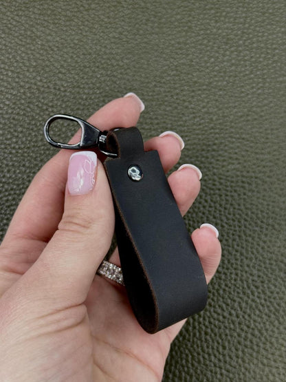 A hand with pink manicured nails holds a black leather keychain strap with a silver metal lobster clasp against a textured olive green background.