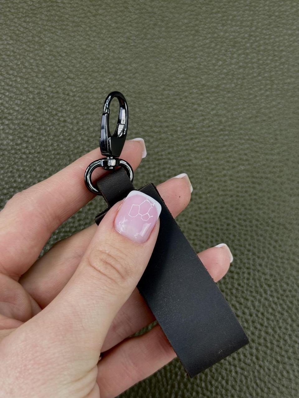 A close-up of a hand holding a black leather keychain with a black metal carabiner against a textured olive green background.