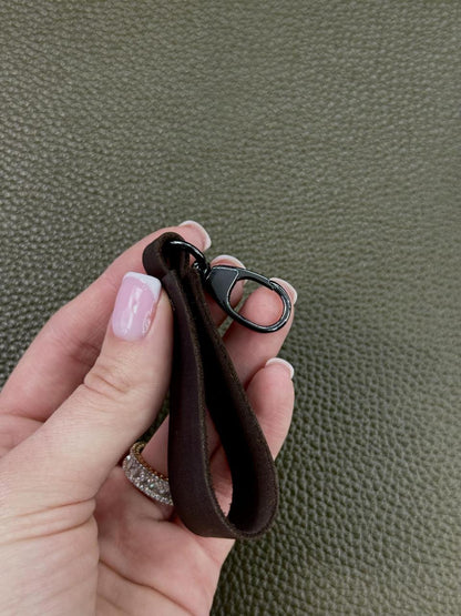 A hand holding a dark brown fabric wrist strap keychain with a black metal lobster clasp, set against a textured olive green background.
