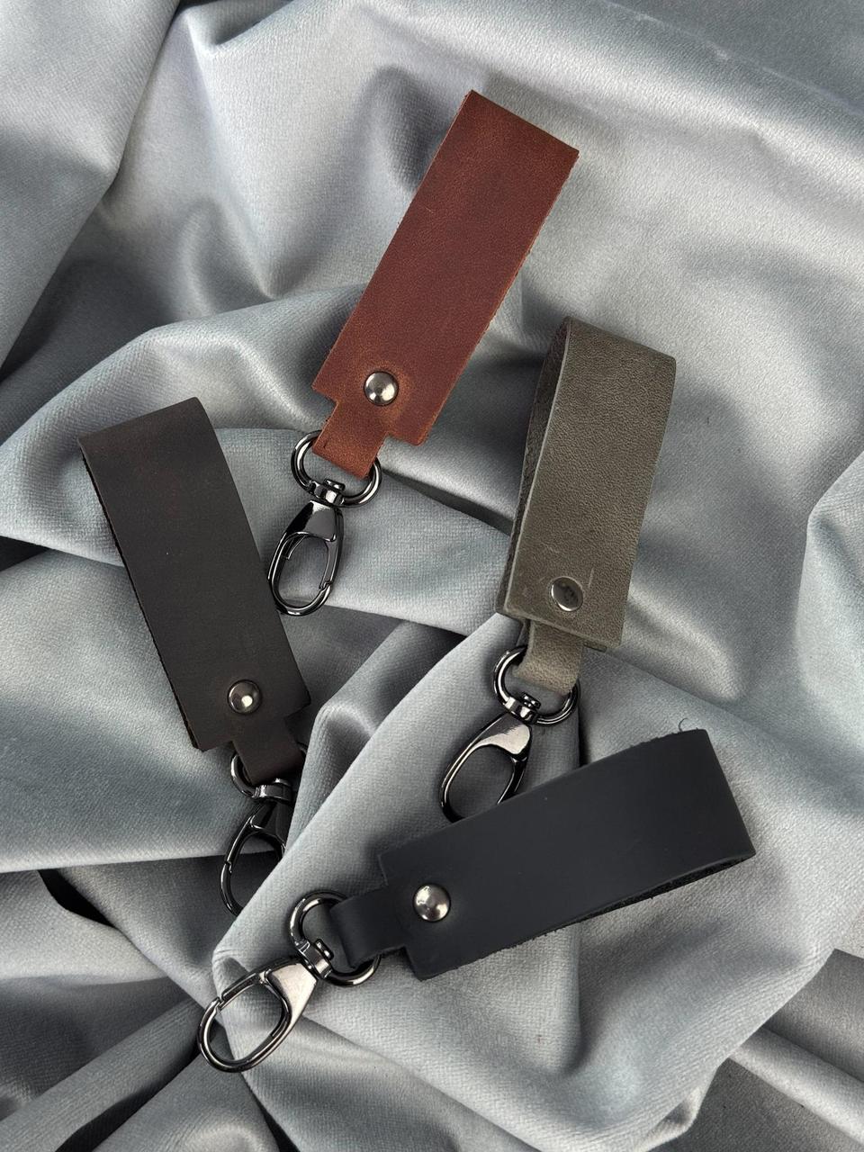 Four leather keychain tags in brown, black, and gray, each with a metal key ring and clasp, arranged on a draped light gray satin fabric background.
