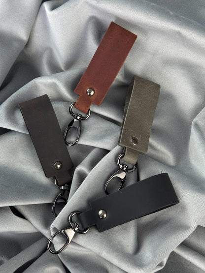 Four leather keychain tags in brown, black, and gray, each with a metal key ring and clasp, arranged on a draped light gray satin fabric background.