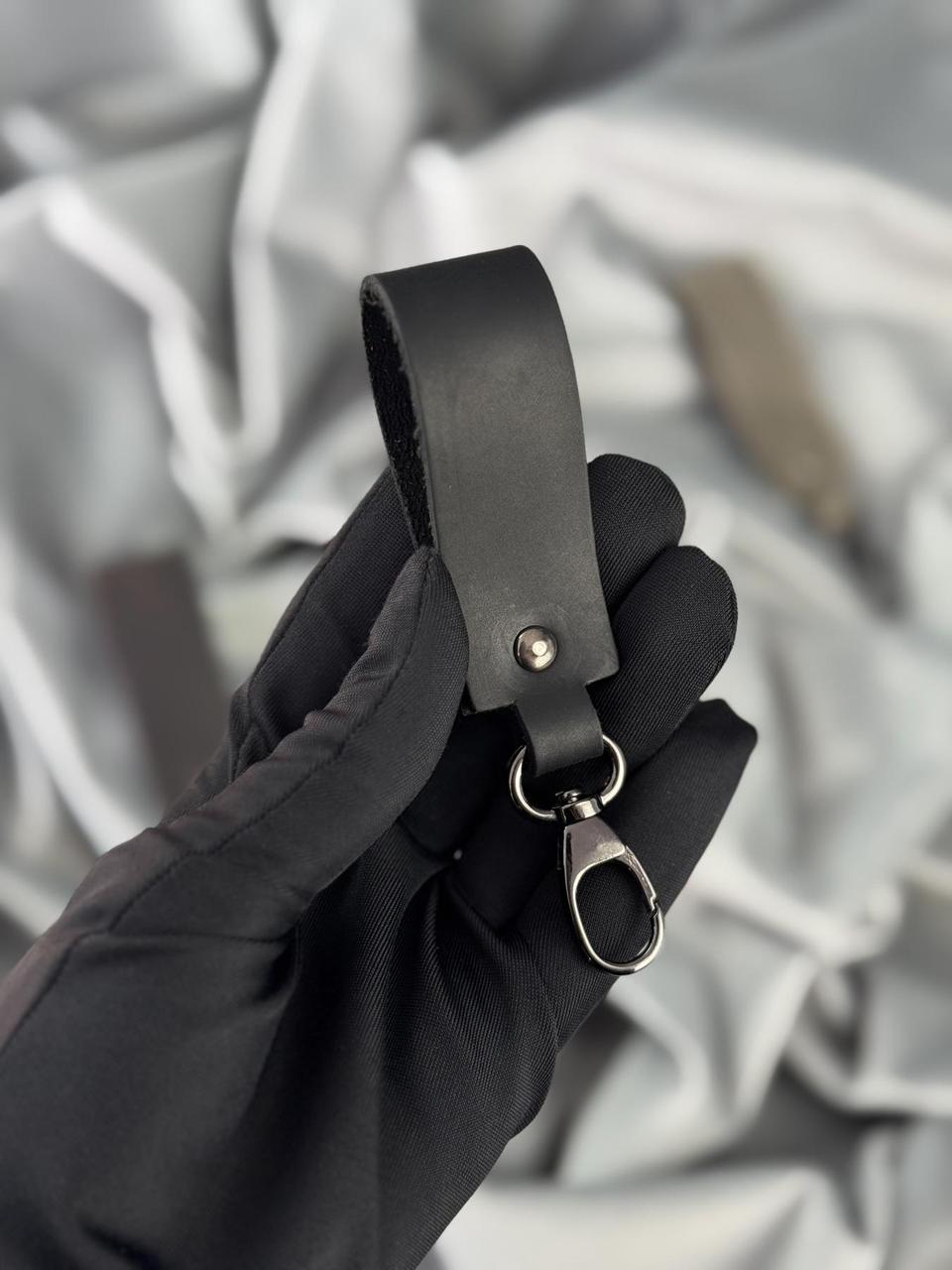 A black leather key fob or wrist strap with a silver metal snap button, ring, and lobster clasp, held by a hand wearing a black glove against a soft gray fabric background.