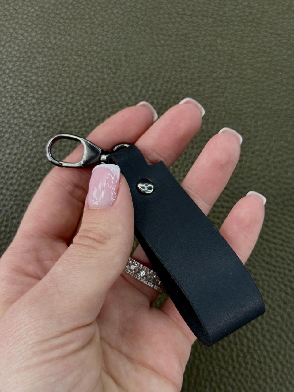 A close-up of a hand holding a black leather keychain strap with a silver metal lobster clasp. The hand has light pink manicured nails and a silver ring with clear stones, set against a textured olive green background.
