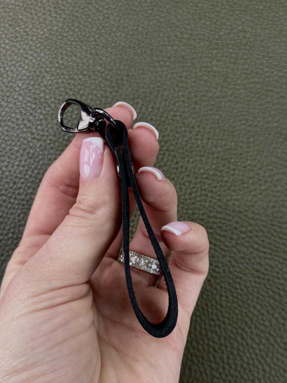 A close-up of a hand holding a black fabric wrist strap keychain with a silver-tone lobster clasp, set against a textured olive green background.