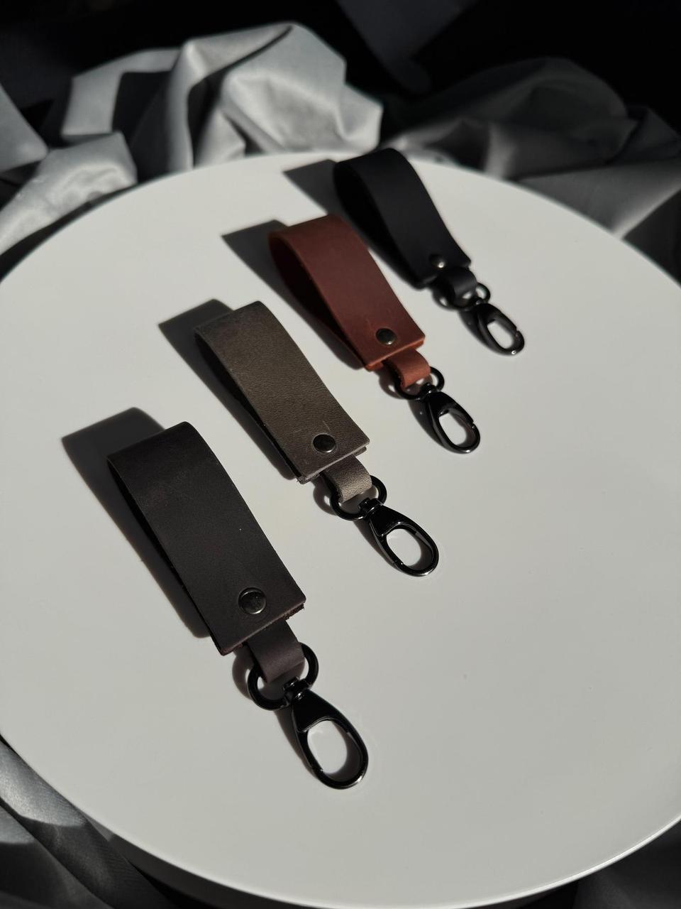 Three leather keychain clips in black, brown, and dark gray, arranged diagonally on a white circular surface with draped gray fabric in the background.