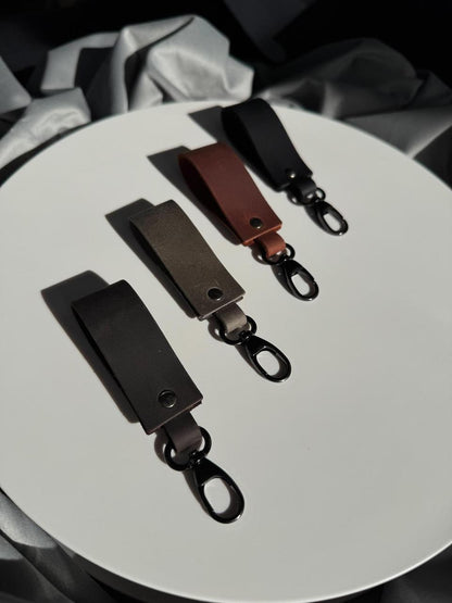 Three leather keychain clips in black, brown, and dark gray, arranged diagonally on a white circular surface with draped gray fabric in the background.