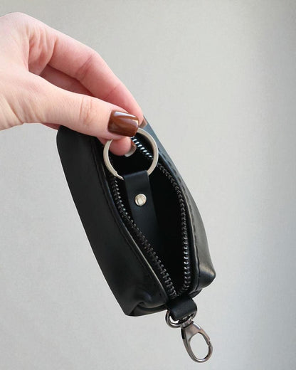 A hand with brown nail polish holds open a small black leather zip pouch with a silver metal key ring and clip, against a plain light gray background.