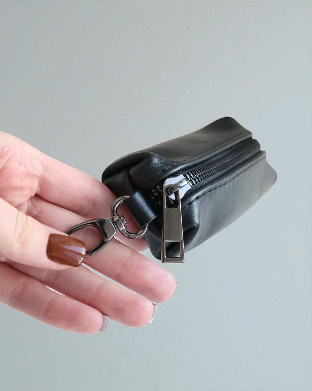 A hand with brown nail polish holds a small black zippered pouch with a silver metal zipper pull and key ring attachment against a plain light gray background.