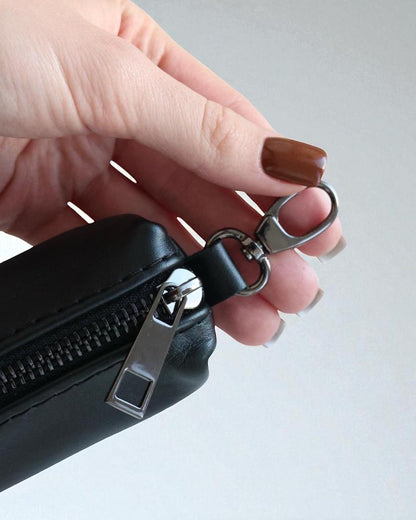 A close-up of a hand with brown nail polish holding a black faux-leather zippered pouch attached to a silver metal carabiner clip, against a light gray background.
