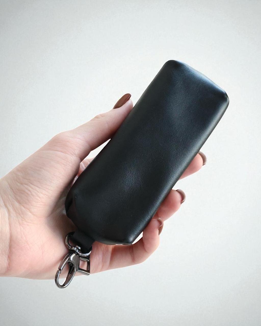 A hand holding a black leather key case with a silver metal keychain ring, set against a plain light gray background.