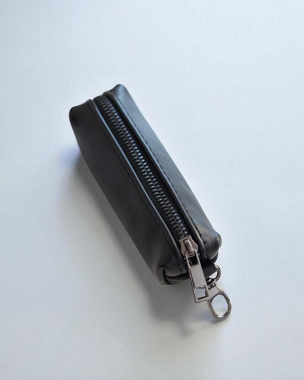 A small black leather zippered pouch with a silver metal zipper pull and key ring, photographed on a light gray background.