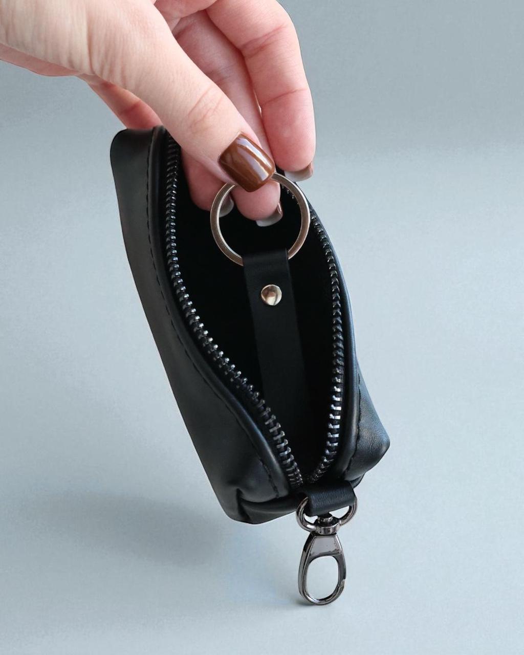 A hand with brown nail polish holds a small black leather zip pouch with a silver metal key ring and clip against a light gray background.