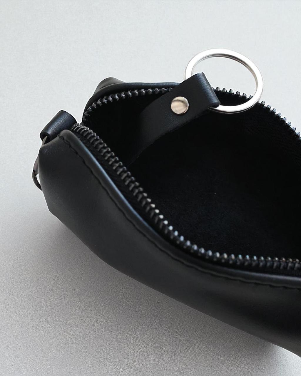 Close-up of a black leather zippered pouch with a silver metal key ring and a small silver rivet, shown open on a light gray background.