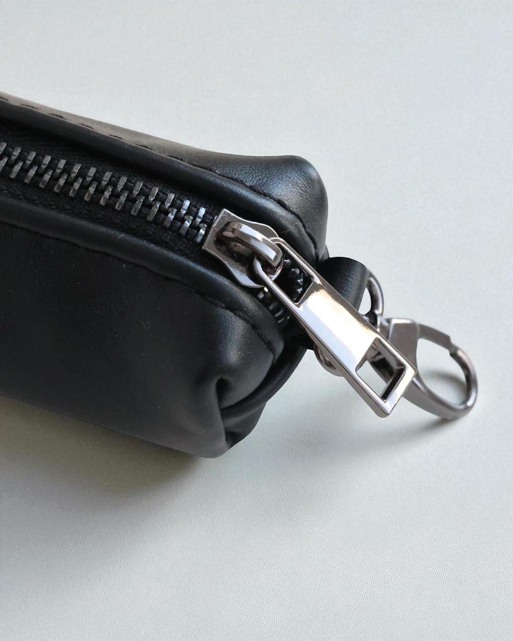 Close-up of a black leather zippered pouch with a silver metal zipper pull and keychain ring, set against a light gray background.