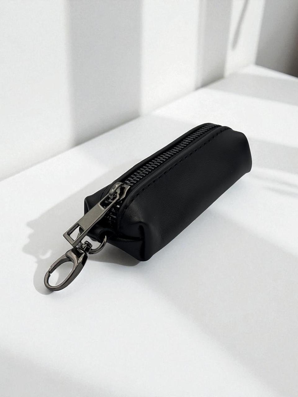 A small black zippered pouch with a silver metal key ring clasp, resting on a white surface with soft shadows.