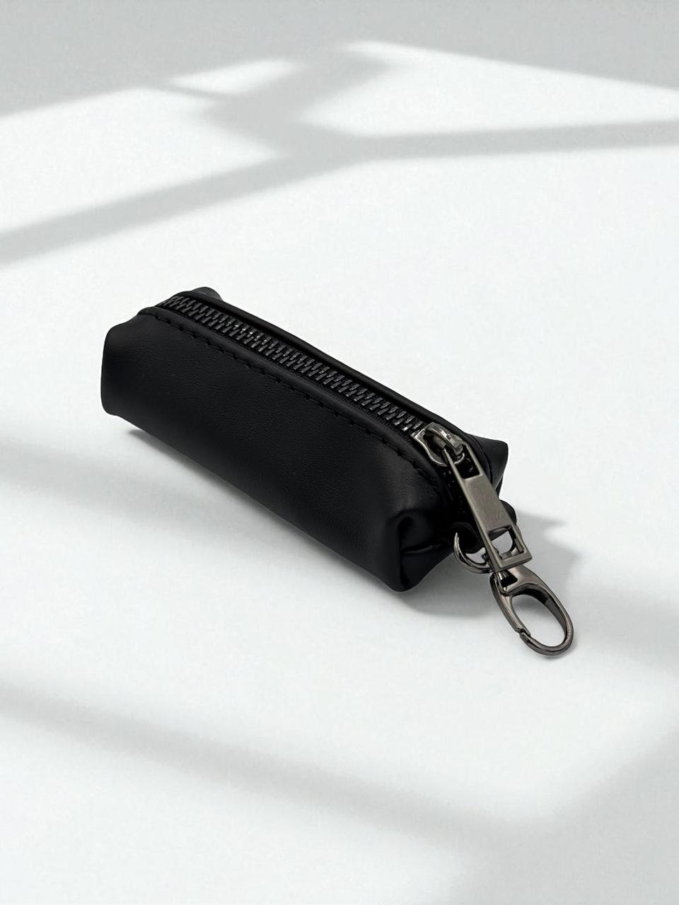 A small black zippered pouch with a silver metal clasp and key ring, photographed on a white surface with soft shadows.