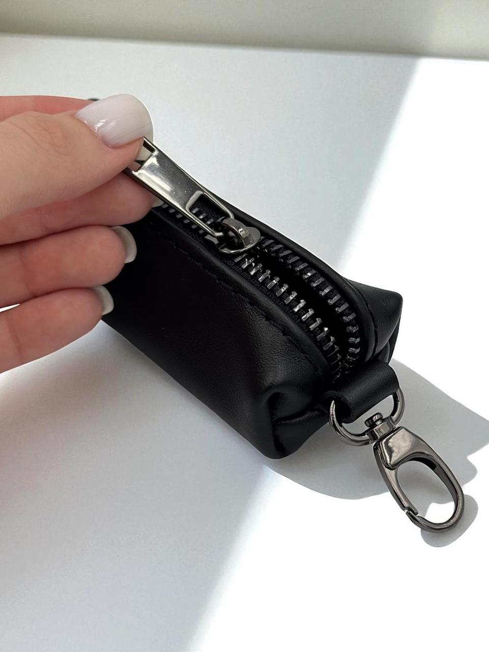 A hand with light pink manicured nails holds a small black zippered pouch with a silver metal clasp and keychain ring, set against a bright white background with soft shadows.