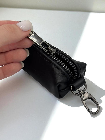 A hand with light pink manicured nails holds a small black zippered pouch with a silver metal clasp and keychain ring, set against a bright white background with soft shadows.