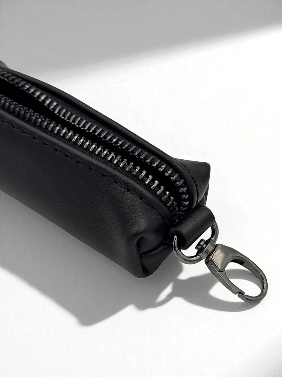 A close-up of a black leather zippered pouch with a silver metal swivel clasp, shown on a white surface with soft diagonal shadows.