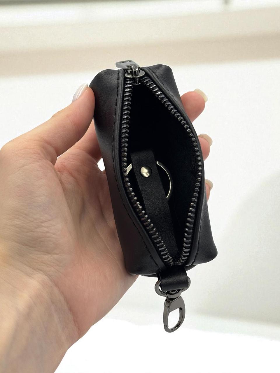 A hand holding a small black zippered pouch with a black interior and silver-tone zipper hardware against a light background.
