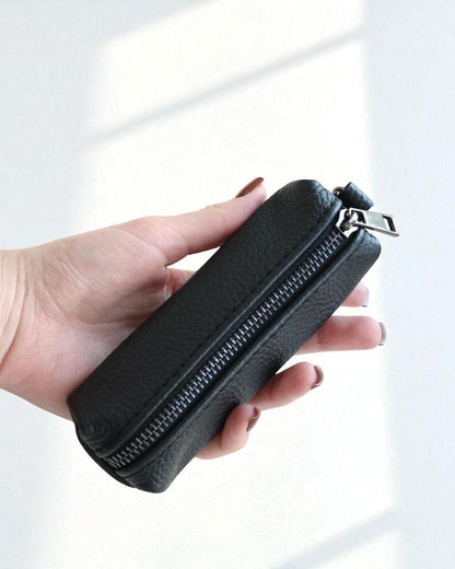 A hand holding a small black pebbled leather zippered pouch against a bright white background with soft window shadows.