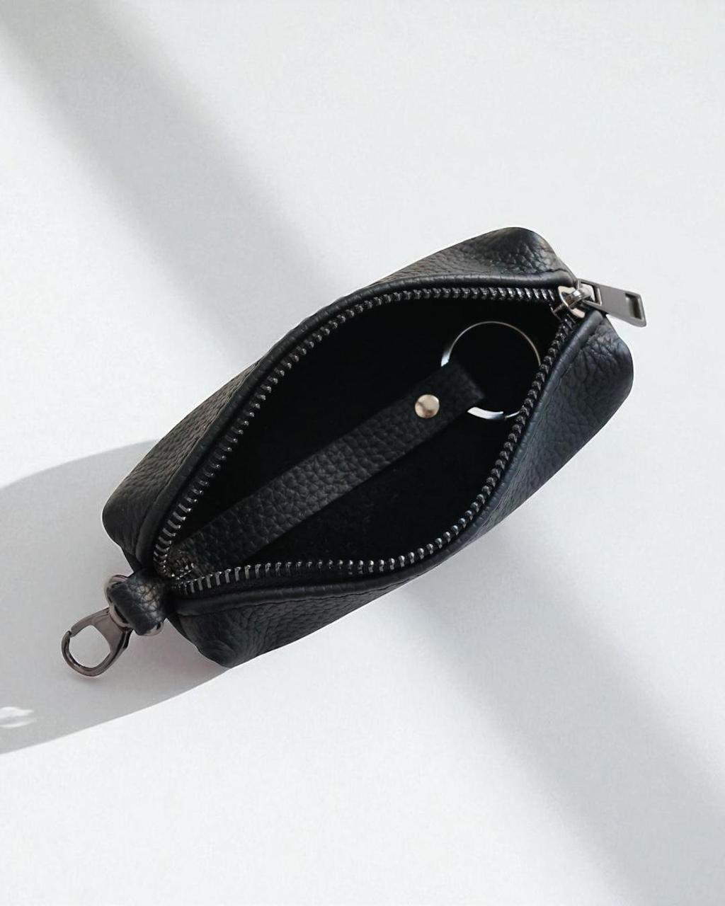 A black textured leather zippered pouch shown open, revealing a black interior and a silver-tone key ring, photographed on a white surface with soft diagonal shadows.