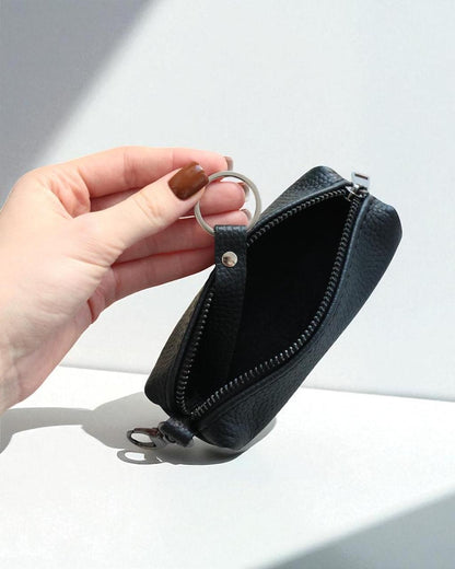 A hand with brown nail polish holds open a small black pebbled leather zip pouch with a silver metal key ring attachment, set against a clean white background with soft shadows.