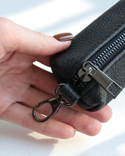 A close-up of a hand holding a black pebbled leather zippered pouch with a silver-tone metal zipper pull and black keychain ring attachment, set against a bright white background.