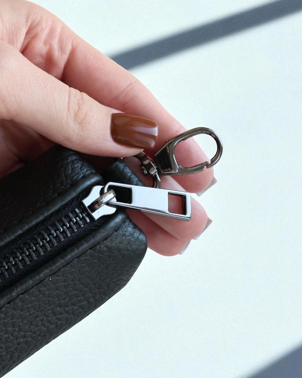 A close-up of a hand with brown manicured nails holding a black textured leather zippered pouch, showing a silver metal zipper pull and keychain ring attachment.