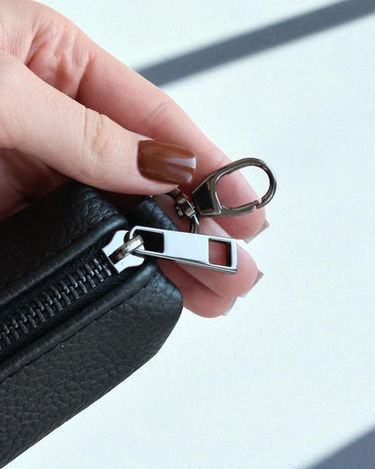 A close-up of a hand with brown manicured nails holding a black textured leather zippered pouch, showing a silver metal zipper pull and keychain ring attachment.