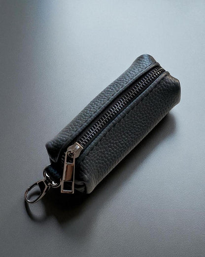 A dark gray pebbled leather zippered pouch with a silver-tone metal zipper pull and keyring attachment, resting on a dark gray textured surface.