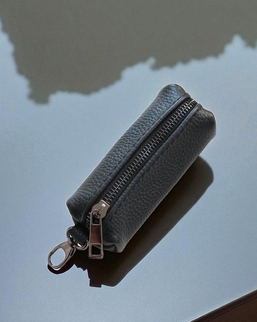 A small pebbled leather zip pouch in a muted blue-gray color with a silver-tone metal zipper and key ring attachment, photographed on a reflective light gray surface.