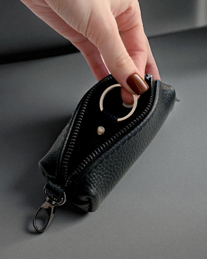 A hand with brown nail polish is shown holding open a small black pebbled leather zip pouch with a silver-tone key ring and carabiner clip, set against a dark gray background.