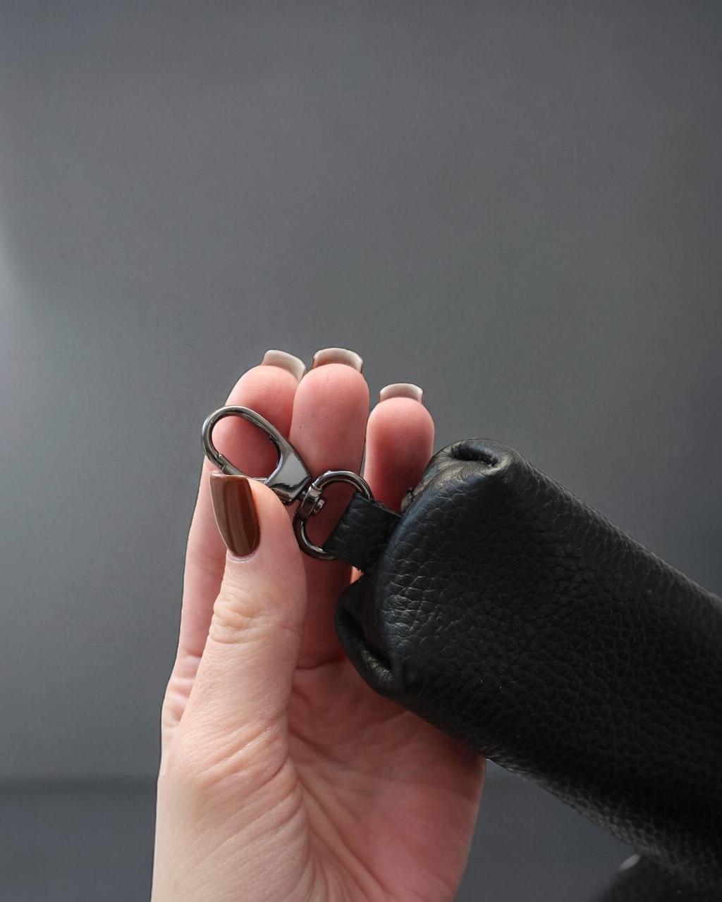 A close-up of a hand holding a black textured leather strap with a silver-toned metal lobster clasp against a dark gray background.