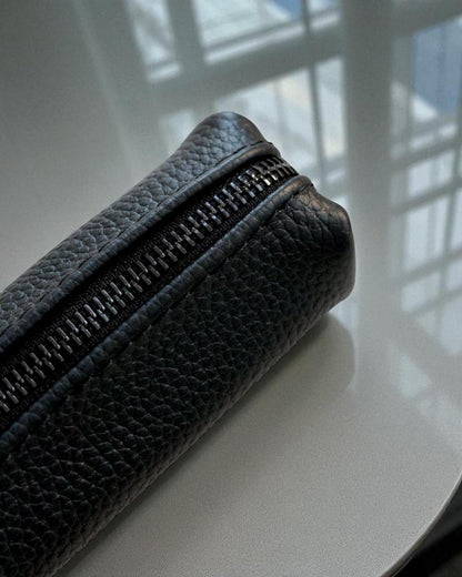 A close-up of a black pebbled leather zippered pouch resting on a glossy white surface with soft window light reflections.