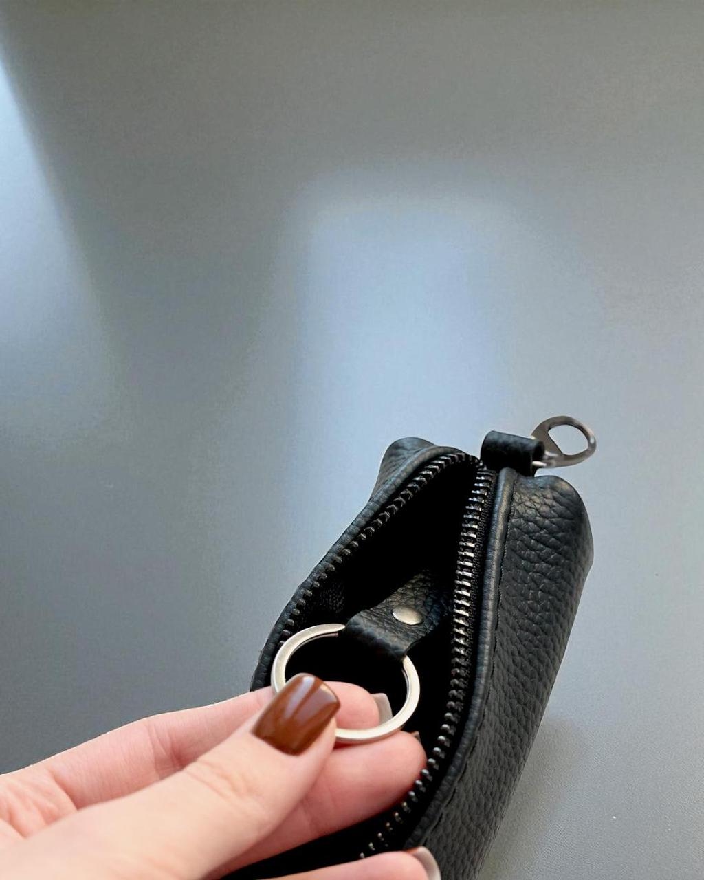 A close-up of a black textured leather zippered pouch with a silver metal key ring, held by a hand with brown manicured nails against a gray background.