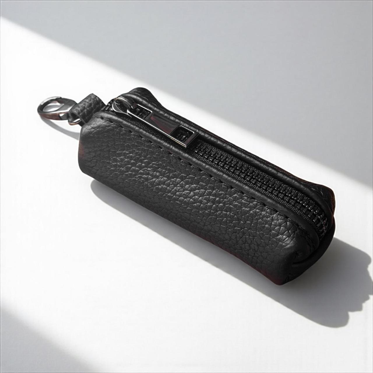 A black pebbled leather zippered pouch with a silver metal key ring, photographed on a white surface with diagonal shadows.