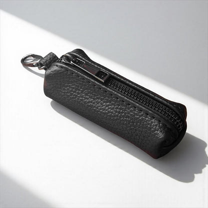 A black pebbled leather zippered pouch with a silver metal key ring, photographed on a white surface with diagonal shadows.