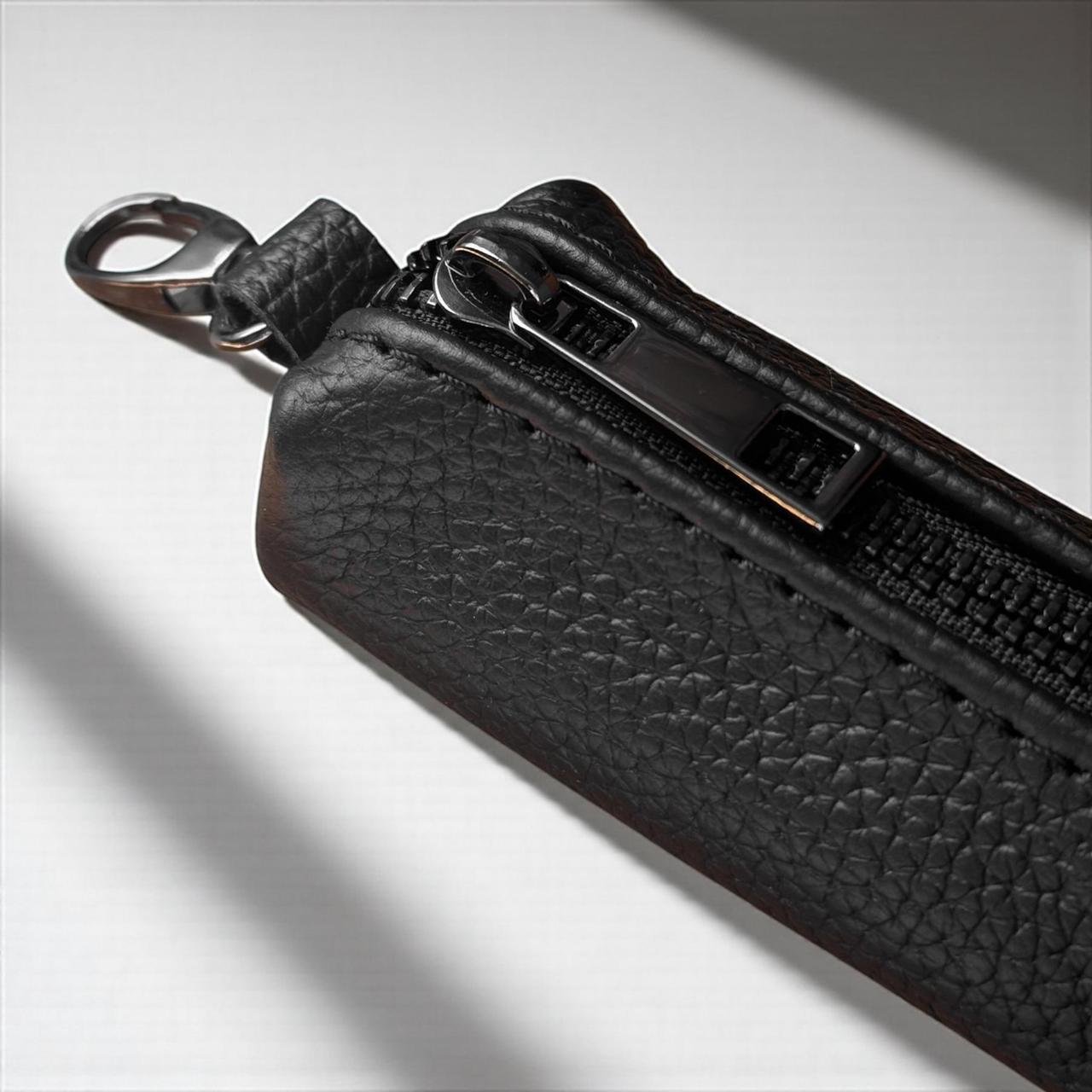 Close-up of a black textured leather zippered pouch with a silver metal clasp and zipper pull, photographed on a light gray background with dramatic shadows.