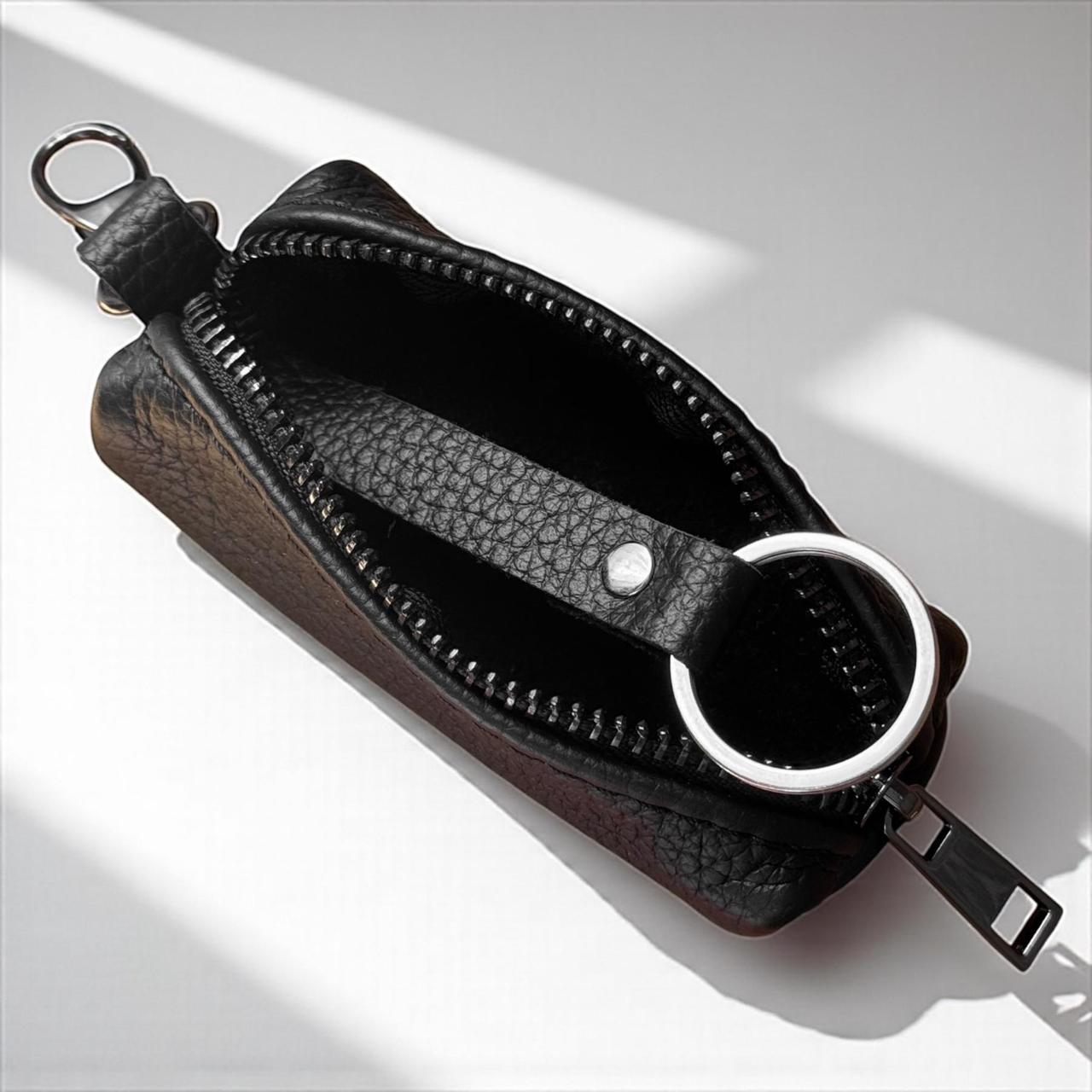 A black textured leather zippered pouch with a silver metal key ring and zipper pull, shown open on a white surface with diagonal shadows.