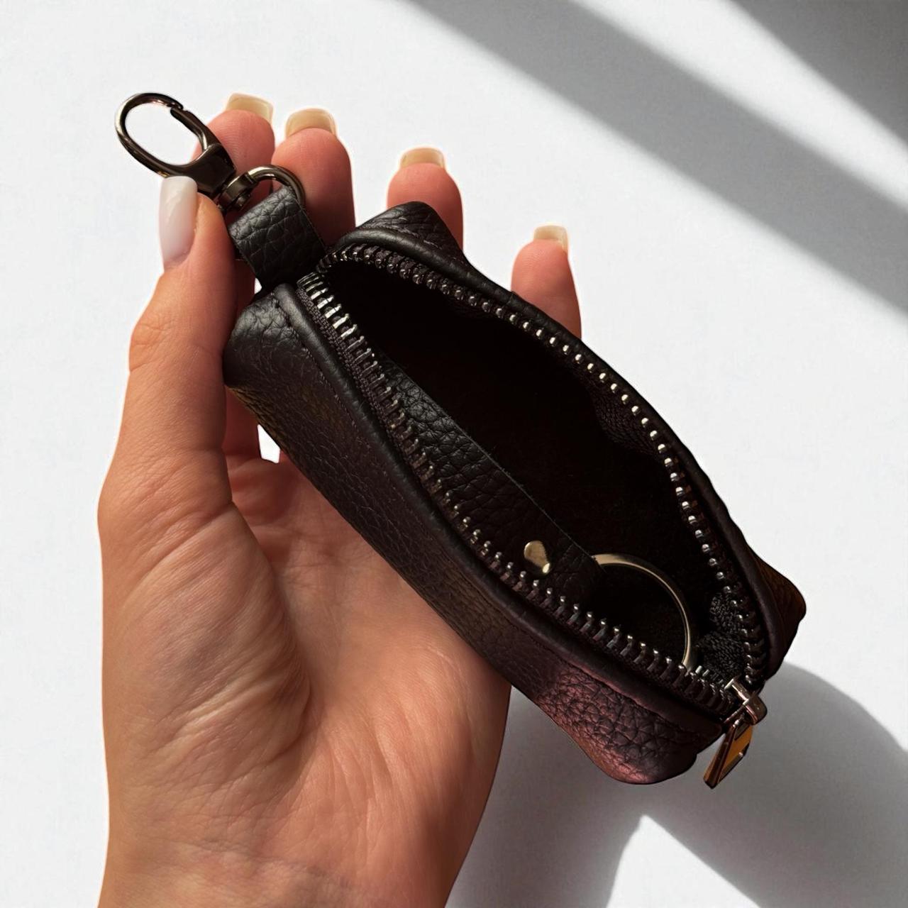 A hand holding a small black pebbled leather zip pouch with a key ring clasp, shown against a white background with soft diagonal shadows.