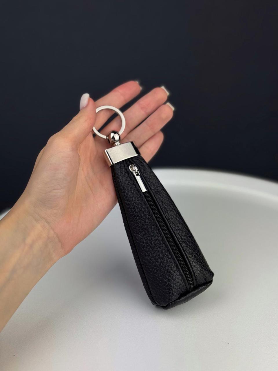 A hand holding a black pebbled leather zippered keychain pouch with silver-tone metal hardware against a dark background.