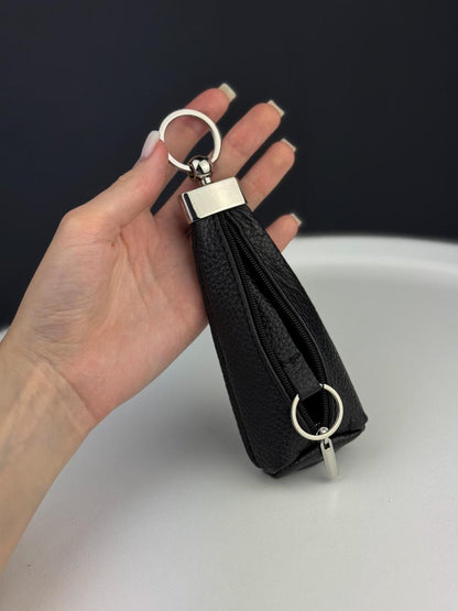 A hand holding a small black pebbled leather zippered pouch keychain with silver-tone metal hardware, including a key ring, rectangular connector, and split key ring.