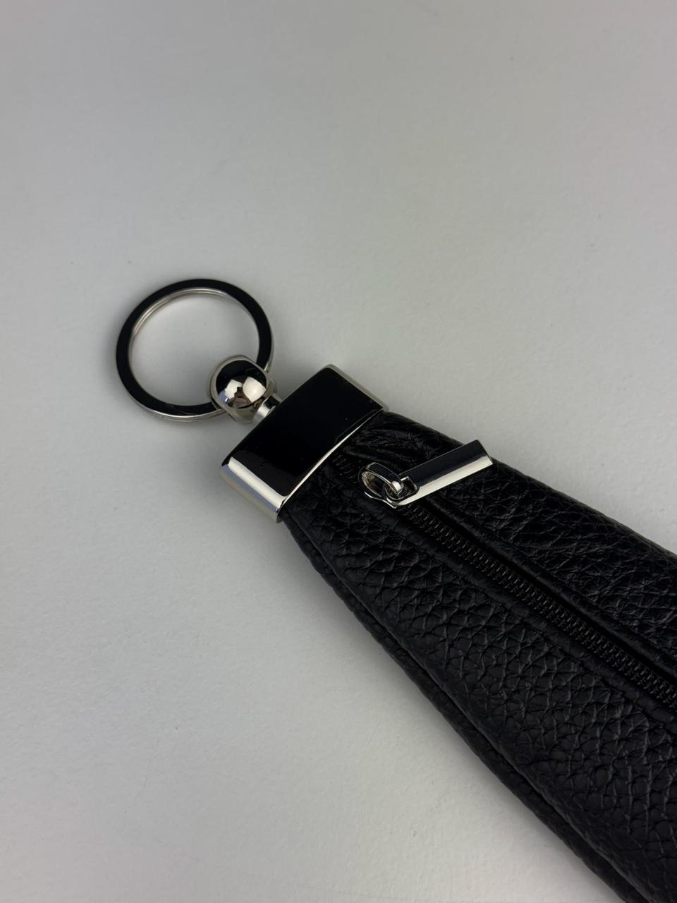 A close-up of a black textured leather wristlet or keychain pouch with a silver-tone metal clasp, zipper pull, and black key ring, set against a plain light gray background.