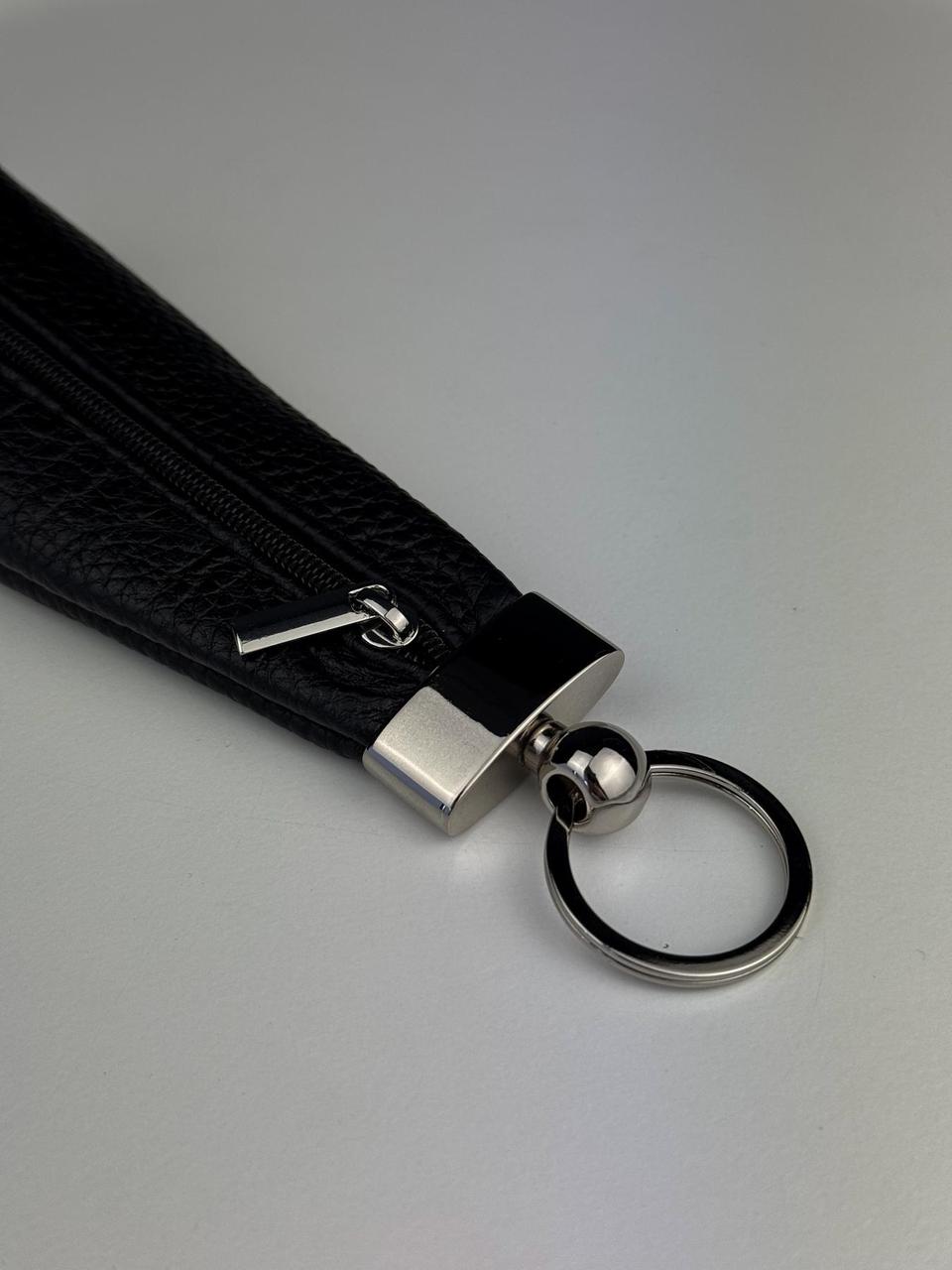 A close-up of a black textured leather wristlet strap with a silver metal clasp, ball connector, and split keyring on a light gray background.