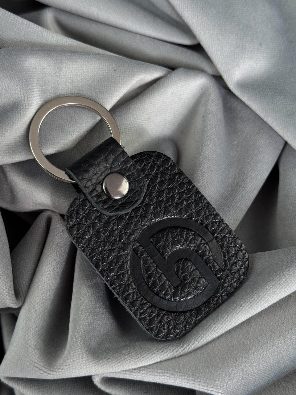 A black textured leather keychain with a silver-tone split ring and a debossed interlocking G logo, resting on draped gray satin fabric.