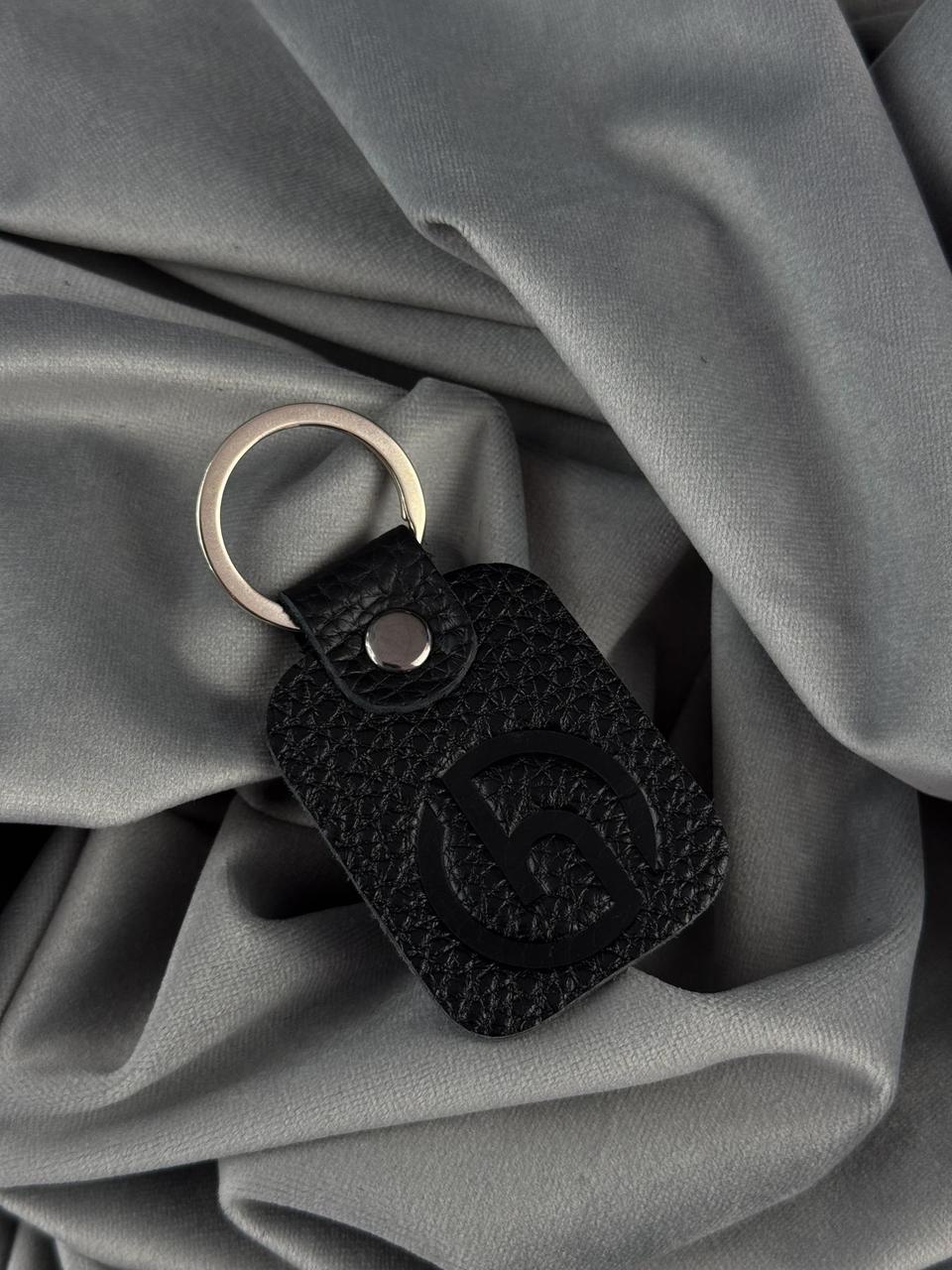 A black textured leather keychain with an embossed logo and a silver metal key ring, resting on draped gray fabric.