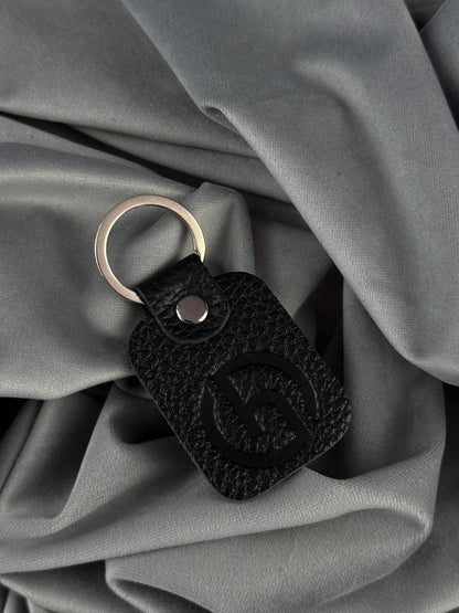 A black textured leather keychain with an embossed logo and a silver metal key ring, resting on draped gray fabric.
