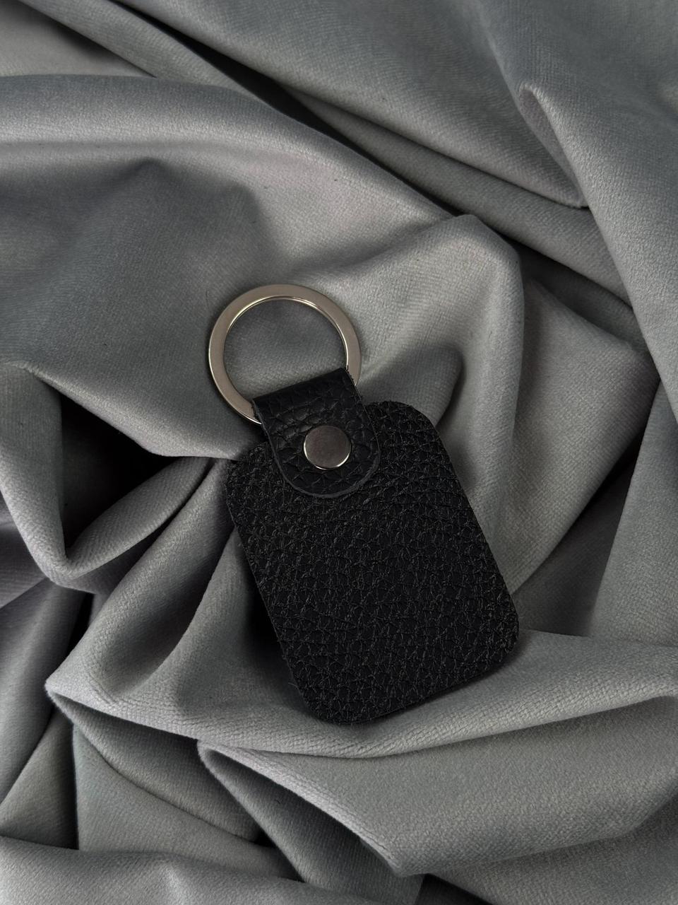 A black textured leather keychain with a silver metal split ring, resting on draped gray fabric.